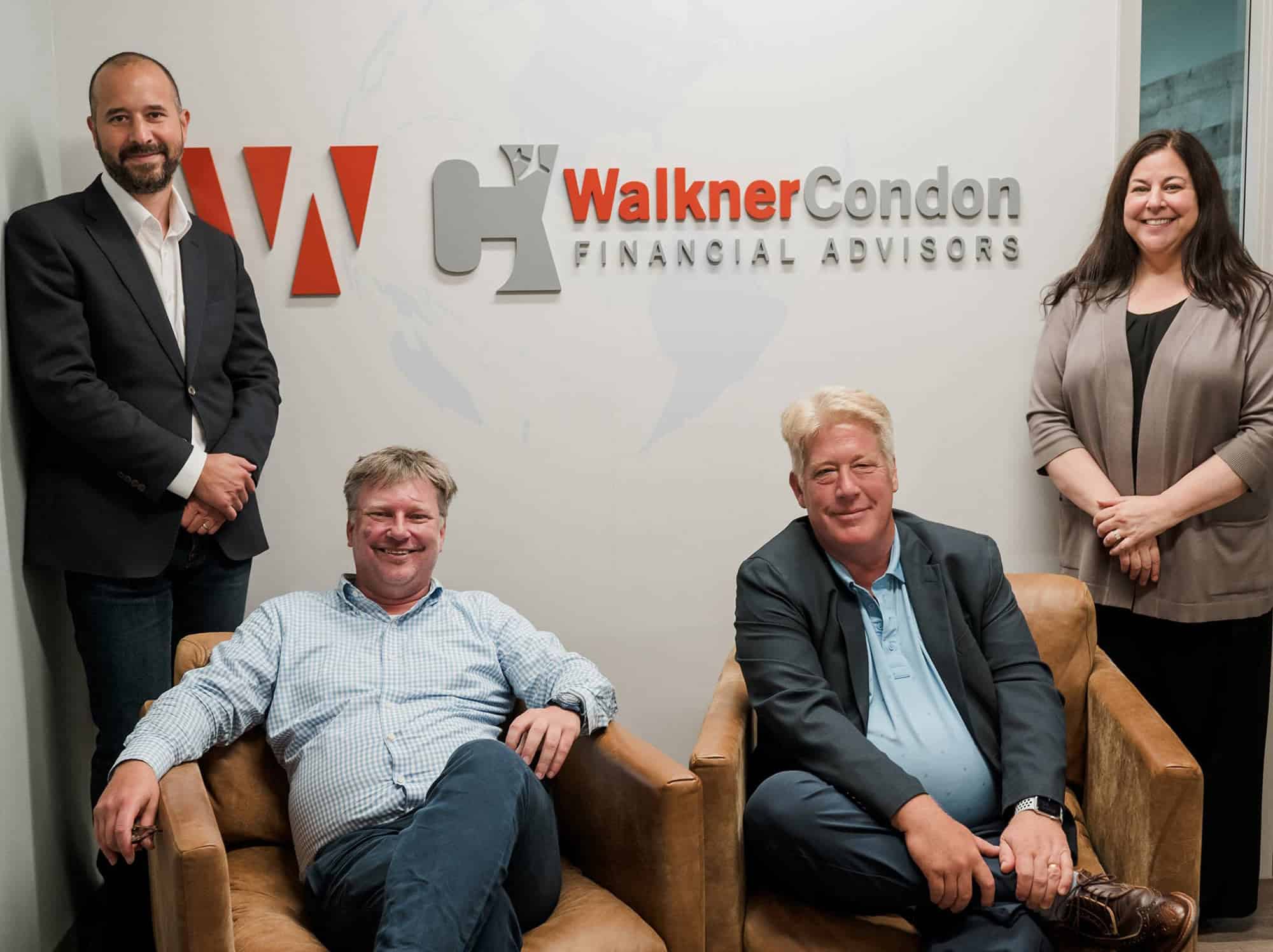Walkner Condon financial advisors Syl, Keith, Stan, and Mary Jo posing together in their office. Syl stands on the left in a dark blazer, Keith is seated on the left in a plaid shirt and dark pants, Stan is seated on the right in a light blue polo and dark blazer, and Mary Jo stands on the right in a gray cardigan. The background features the Walkner Condon logo and a light-colored wall, emphasizing the professional and welcoming environment at Walkner Condon Financial Advisors.