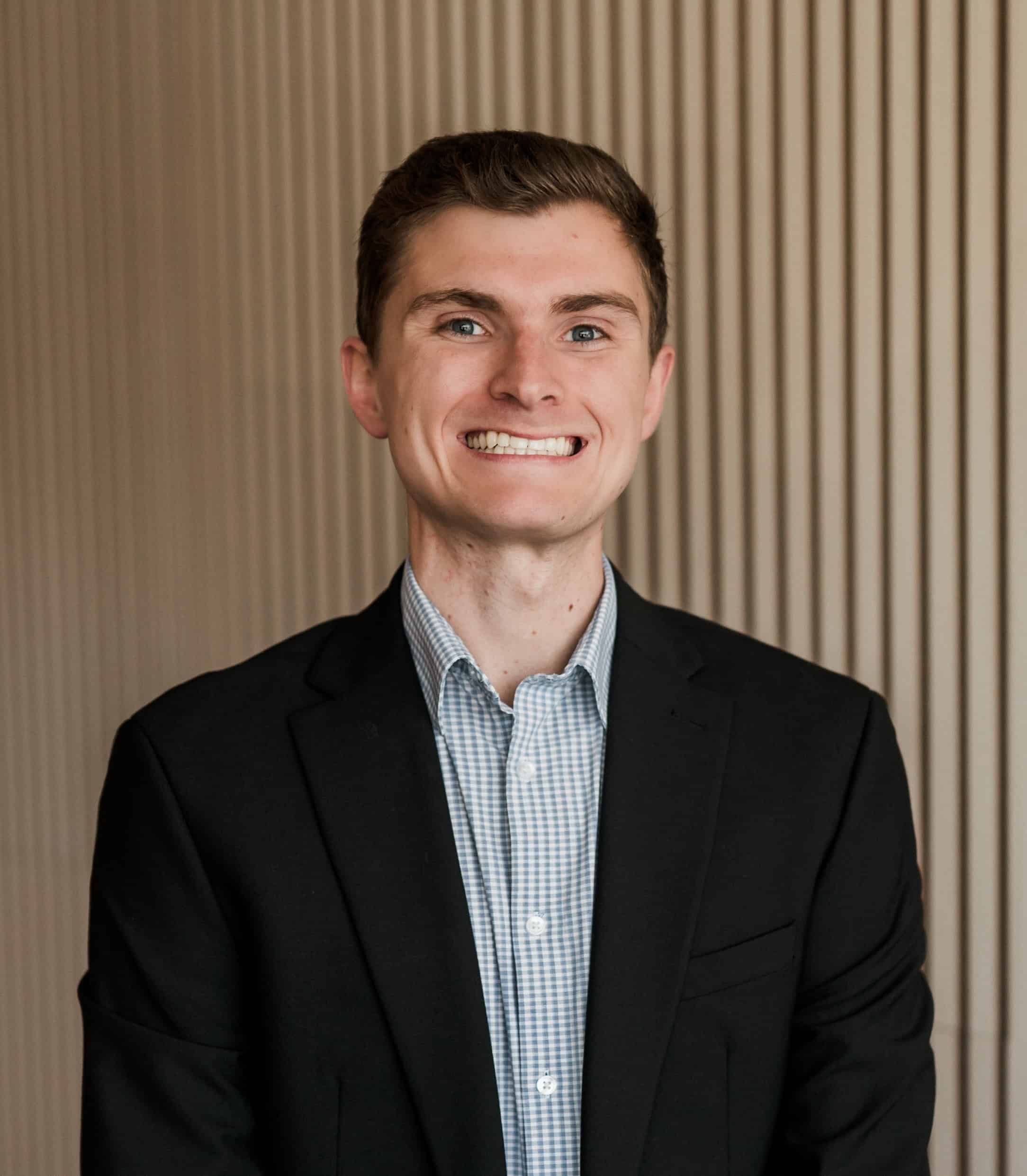 Walkner Condon team member: Young man with short dark hair and a friendly, slight smile. He's wearing a black suit jacket with a light blue and white checkered dress shirt. The background features vertical beige lines, creating a professional office setting.